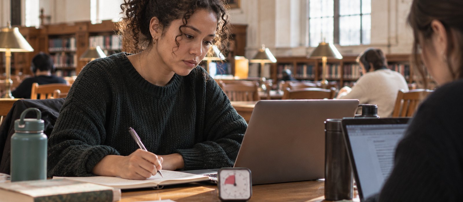 Woman working in a library with a timer, a friend working across the table