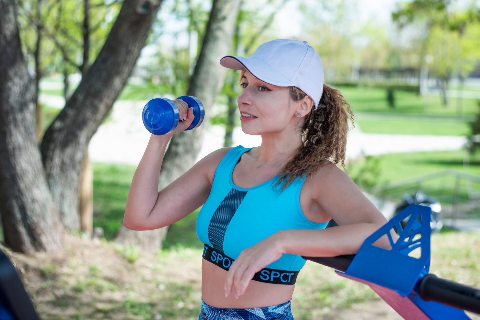 Woman lifting dumbbells in a bright gym with running shoes nearby